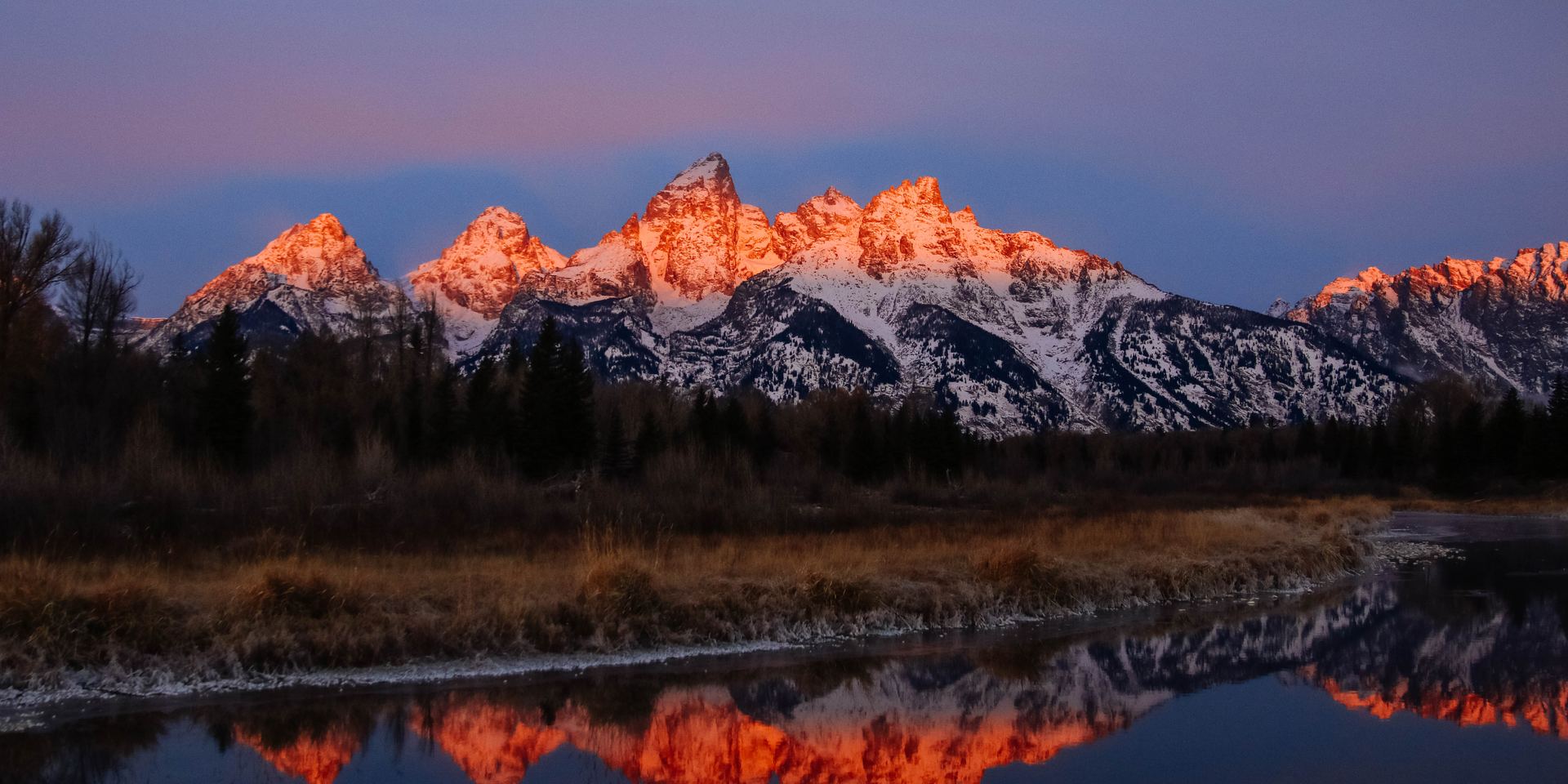The Teton Range at sunrise, its peaks lit orange and reflected in the water at Schwabacher Landing, Jackson Hole, Wyoming